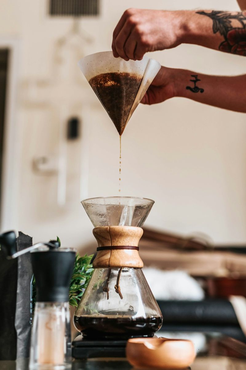 A used paper filter lifted above a Chemex glass brewer, coffee dripping from the cone into the glass