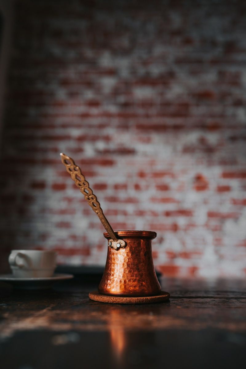 A hammered copper cezve with a long handle beside a white demitasse cup and saucer