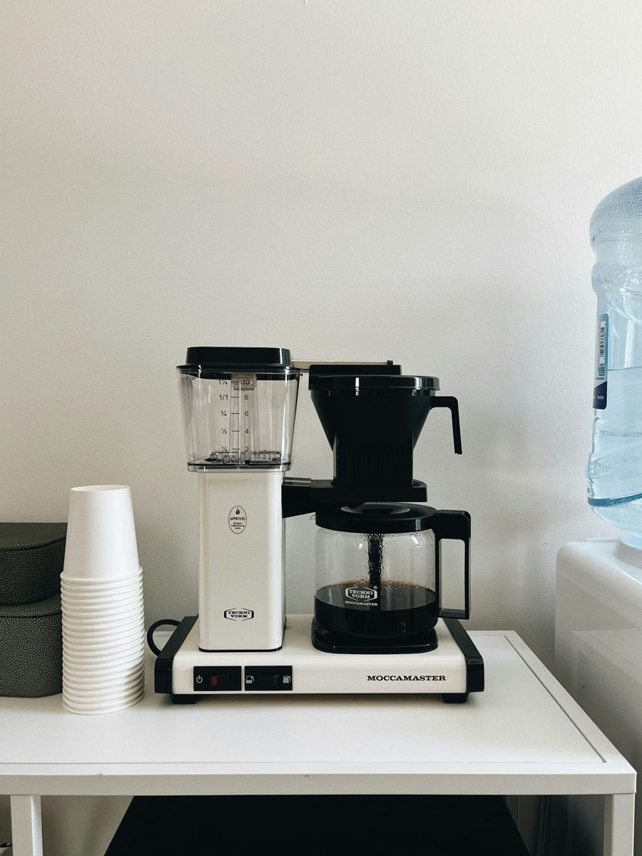 A white Technivorm Moccamaster drip coffee maker mid-brew, coffee streaming into its glass carafe on a white table