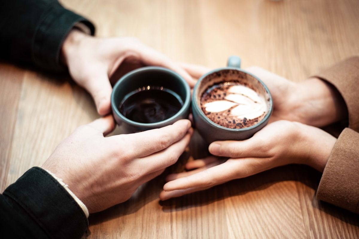 Two people holding ceramic mugs of coffee together across a wooden table