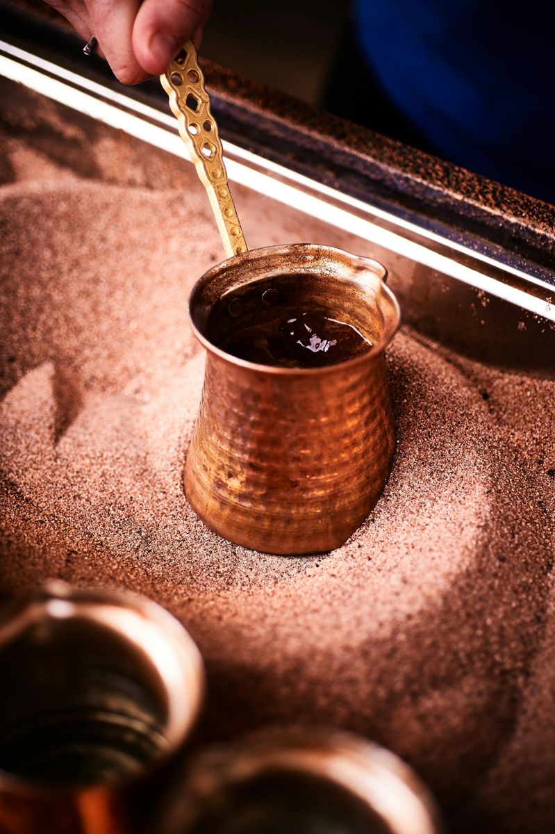 A copper cezve with a long brass handle brewing coffee in a bed of hot sand